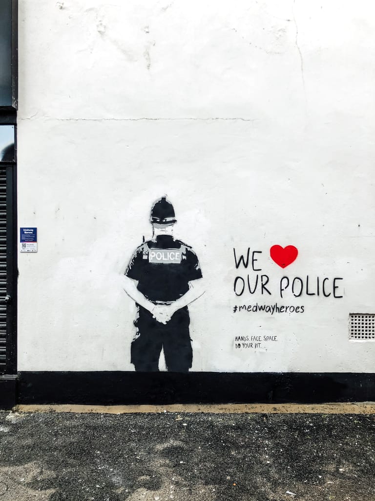 We Heart our Police shows the back of a police officer wearing a custodian helmet and with his arms behind his back. The piece is black and grey stencil art on a white wall. To the right it reads We 'red heart' our police. #medwayheroes Hands. Face. Space. Do your bit. 