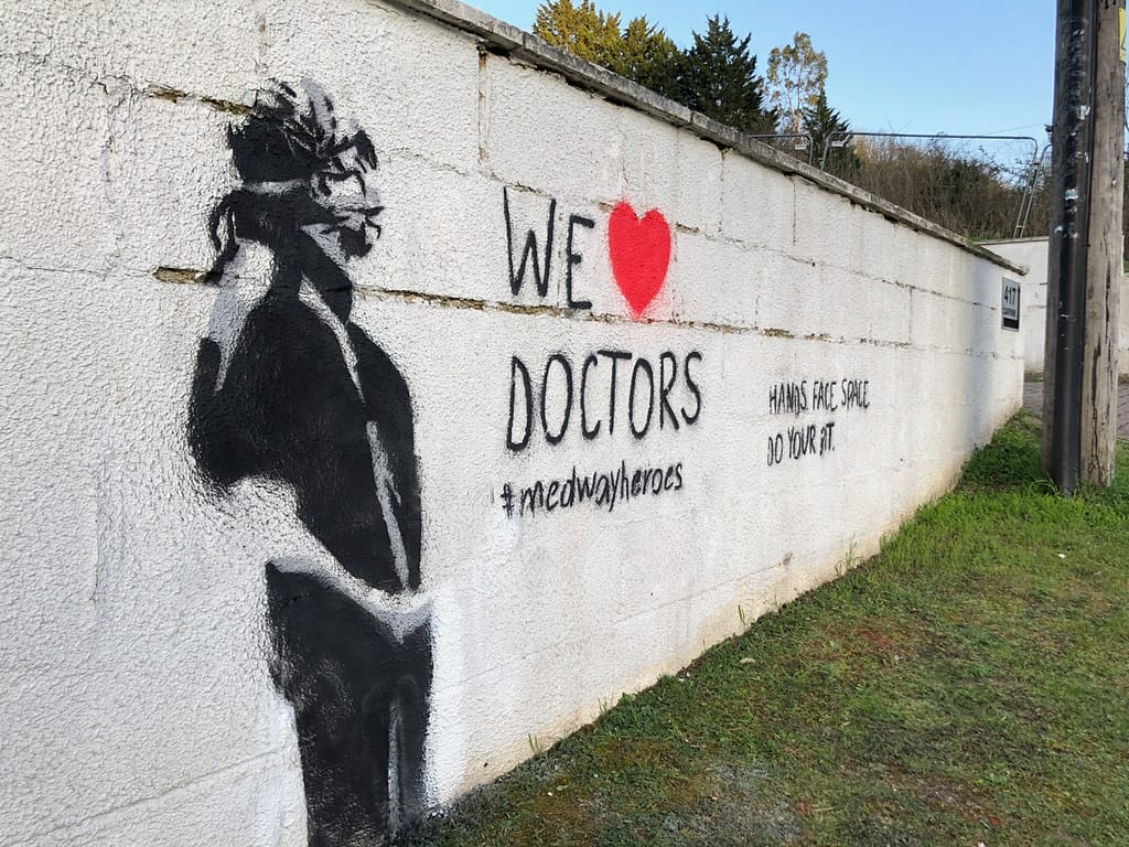'We Heart Doctors' depicts a doctor in PPE sideways looking up. The doctor is in black and grey on a white rough wall. It reads 'We heart doctors' #medwayheroes Hands. Face. Space. to the right along the white fence brick wall. 