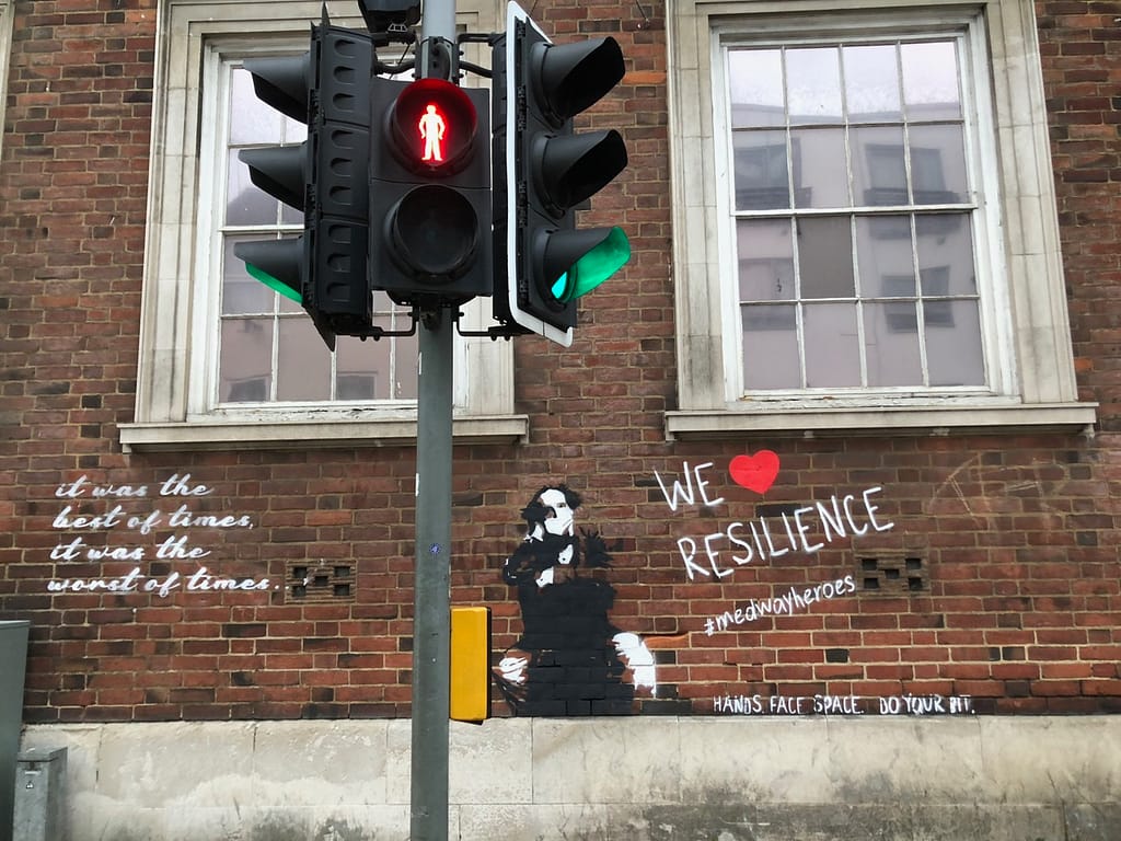'We Heart Resilience' shows Charles Dickens in black and white wearing a mask sitting on a chair on a red brick wall. To the left it reads in cursive writing: it was the best of times, it was the worst of times. To the right it reads 'We Heart Resilience' #medwayheroes Hands. Face. Space. Do your bit.